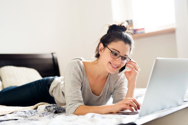 graphicstock-beautiful-young-woman-wearing-eyeglasses-lying-on-bed-working-on-notebook-holding-a-smart-phone-making-a-phone-call-home-office_SO_v1hrfW