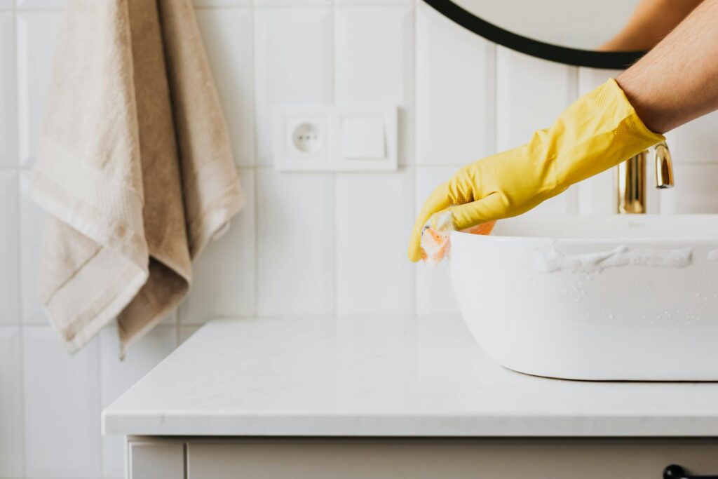 Crop anonymous person wearing yellow latex gloves washing sink in bathroom with orange sponge and cleanser while cleaning contemporary flat