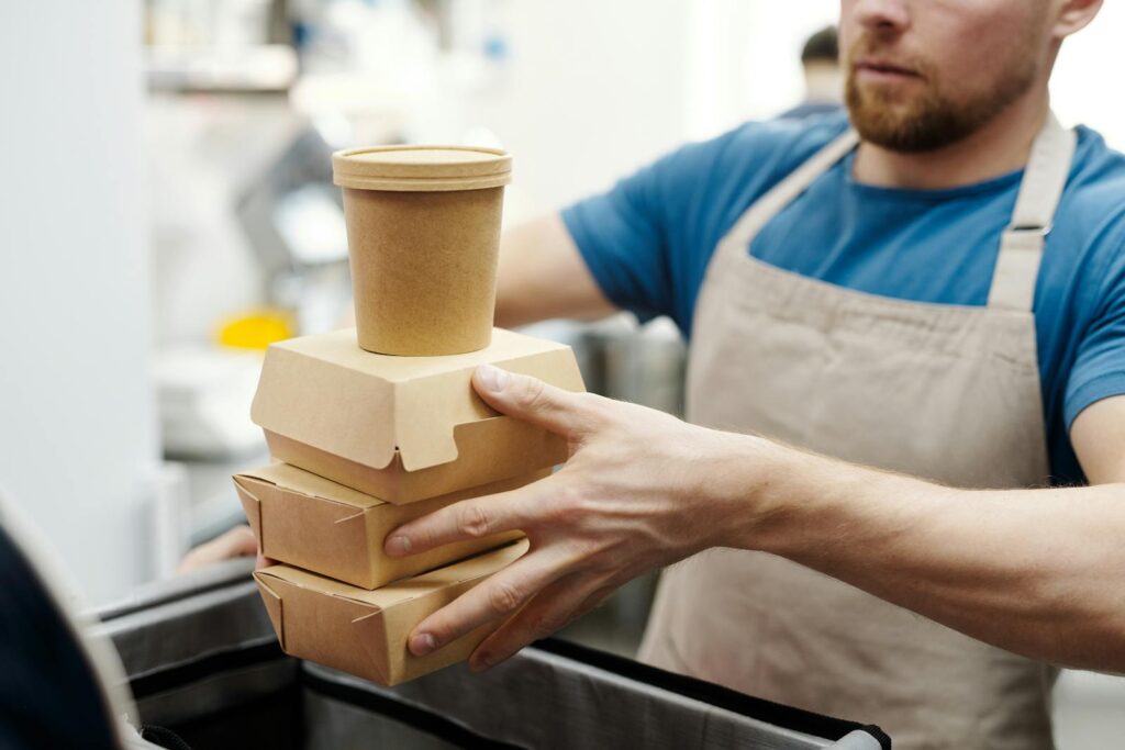 Man wearing apron stacks eco-friendly food containers for takeaway delivery in a kitchen setting.