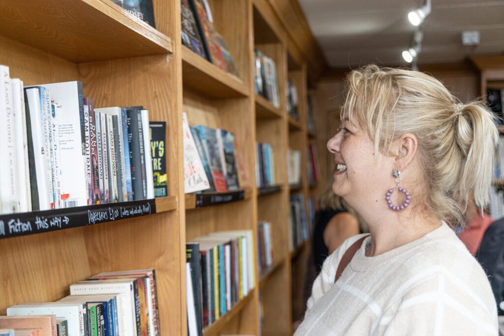 A blonde woman wearing a white top smiling and looking at a bookshelf in a charity shop.