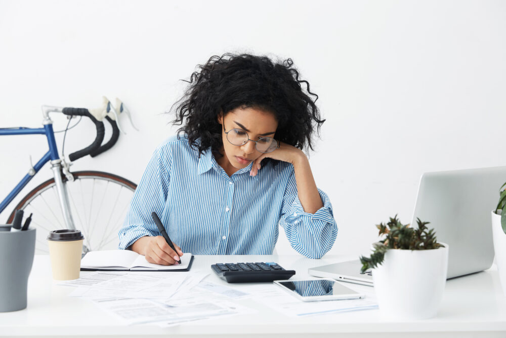 Candid shot of confident hardworking young businesswoman with curly hair sitting at her desk in white office, holding pen while doing accounts by herself, using calculator, laptop and digital tablet