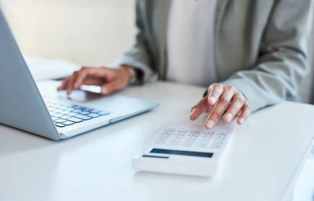 A person wearing a grey cardigan sat at a desk in front of a laptop typing on a calculator.